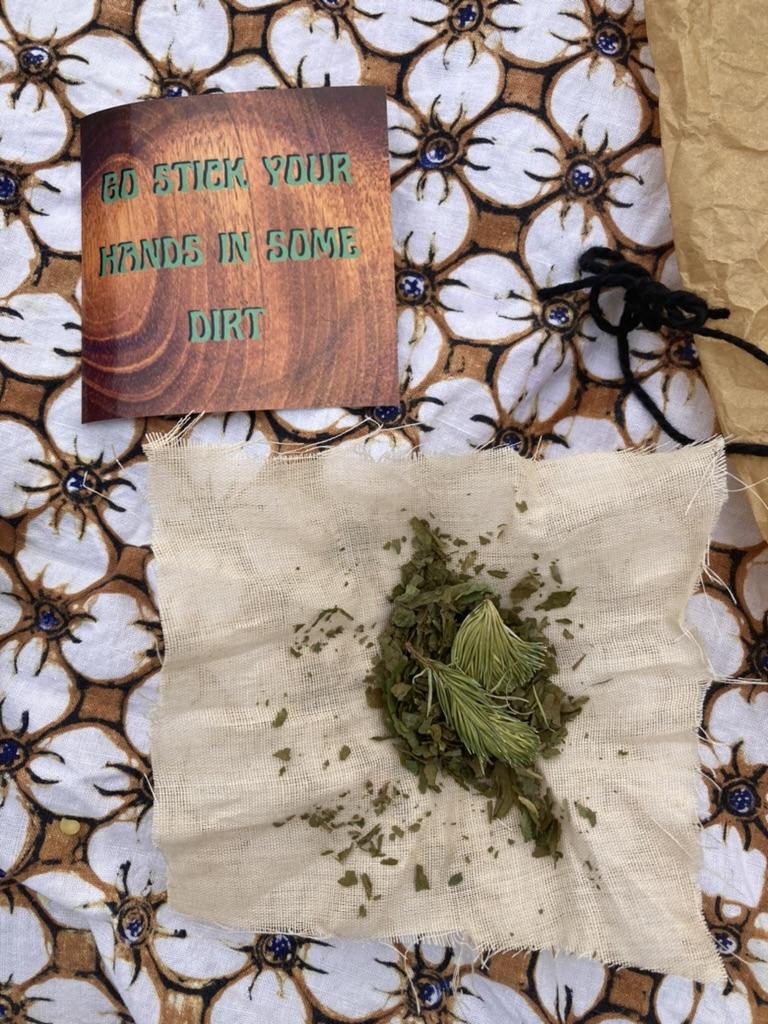 An open cheesecloth sachet holds evergreen needles and other dusty greens, on a table next to a sticker that reads go stick your hands in some dirt.