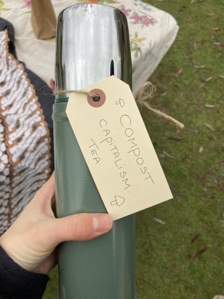 A hand holds a thermos with a tag that reads compost capitalism tea with a small drawing of a mushroom.